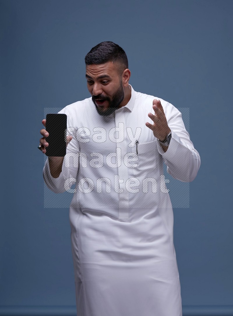 A man Showing his phone in Blue Background wearing Saudi Thob