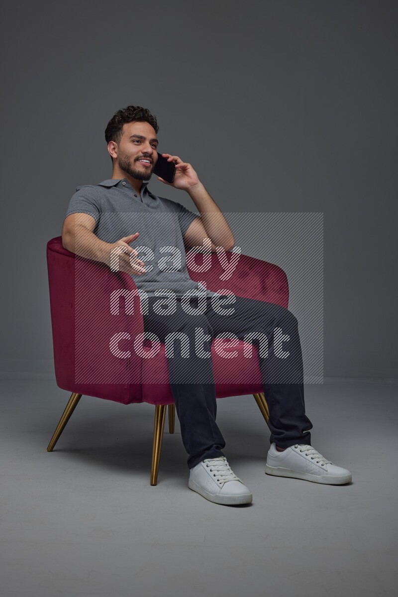 A man wearing casual and talking in his phone while setting on a burgundy chair eye level on a gray background