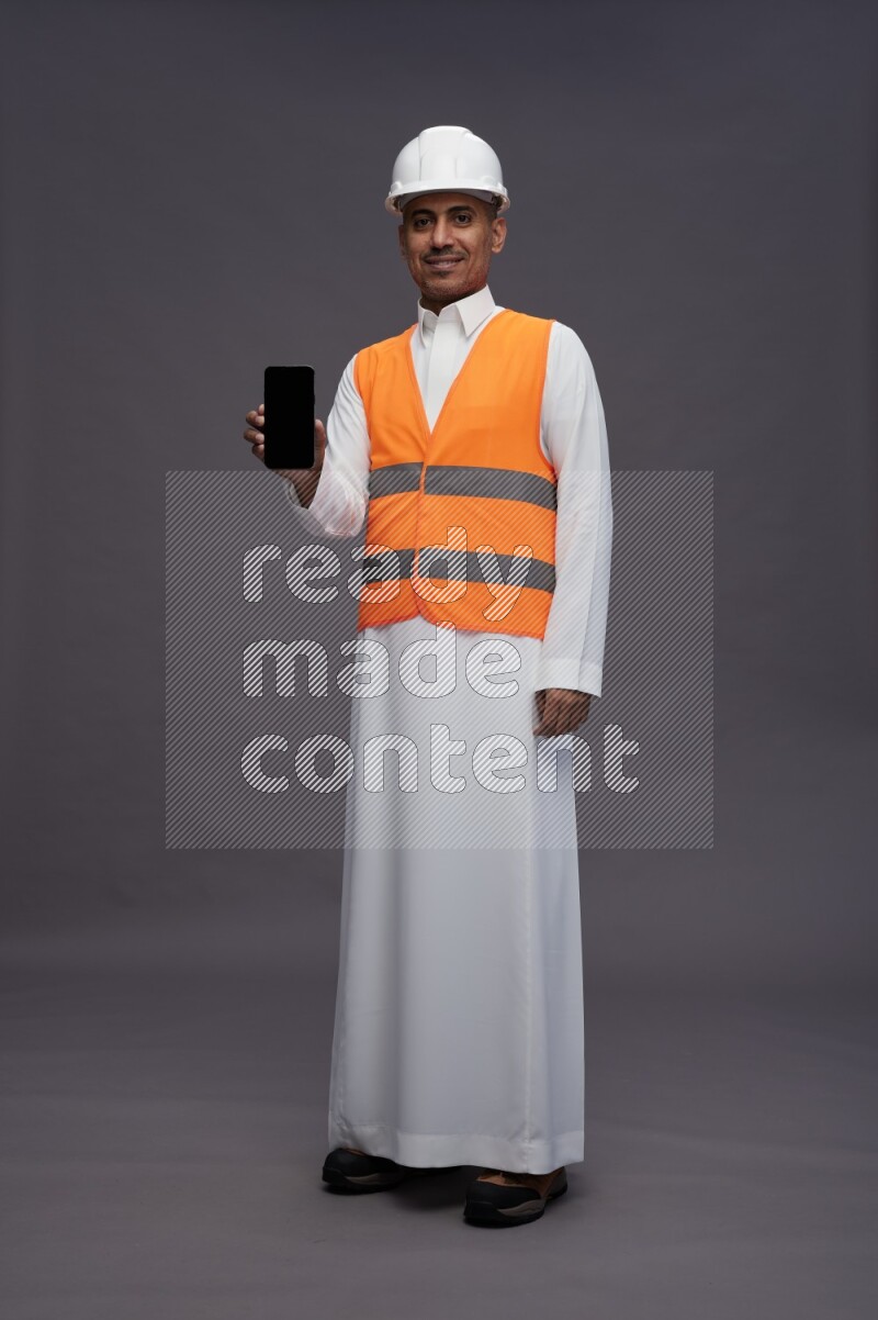Saudi man wearing thob with engineer vest standing showing phone to camera on gray background