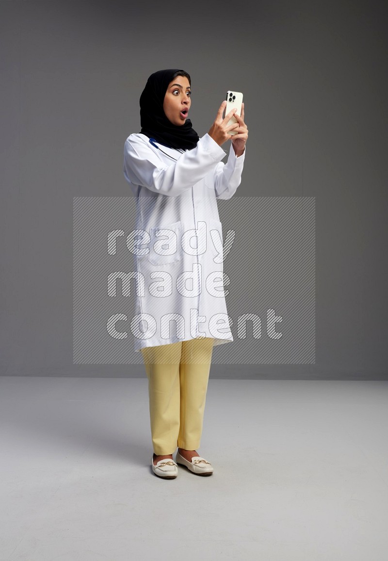 Saudi woman wearing lab coat with stethoscope standing texting on phone on Gray background