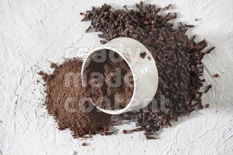 A beige ceramic bowl with cloves powder spilled out of it on a textured white flooring