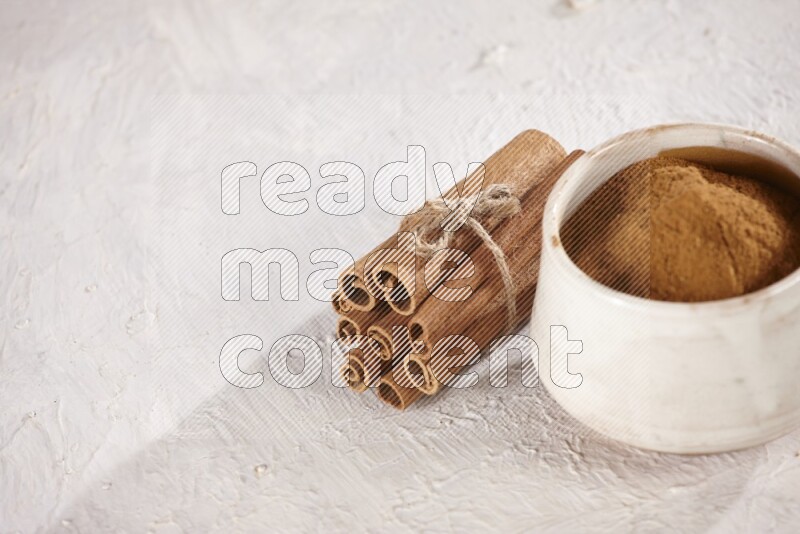 Cinnamon sticks stacked and bounded beside a beige bowl full of cinnamon powder on white background