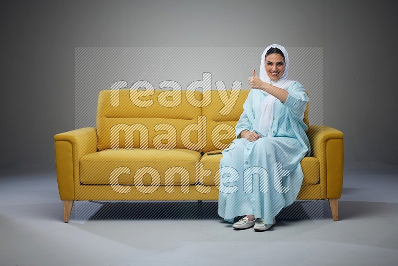 A Saudi woman wearing a light blue Abaya and white head scarf sitting on a yellow sofa and thinking and pointing up eye level on a grey background