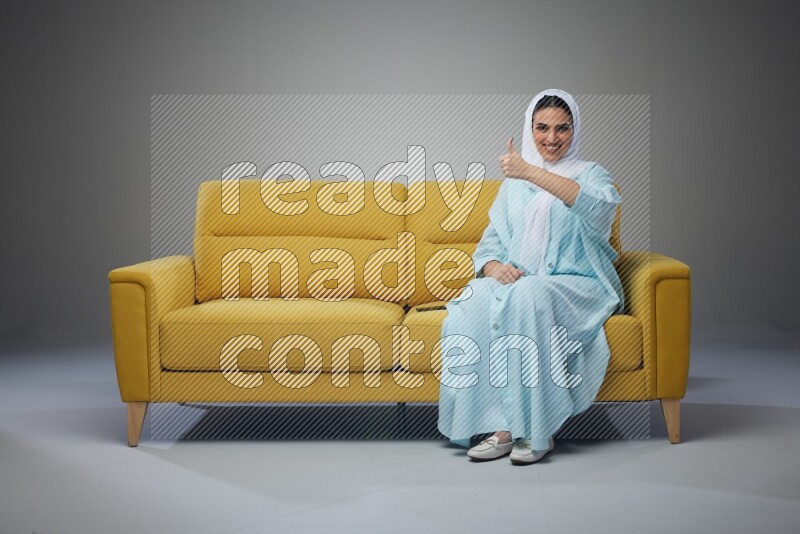 A Saudi woman wearing a light blue Abaya and white head scarf sitting on a yellow sofa and thinking and pointing up eye level on a grey background