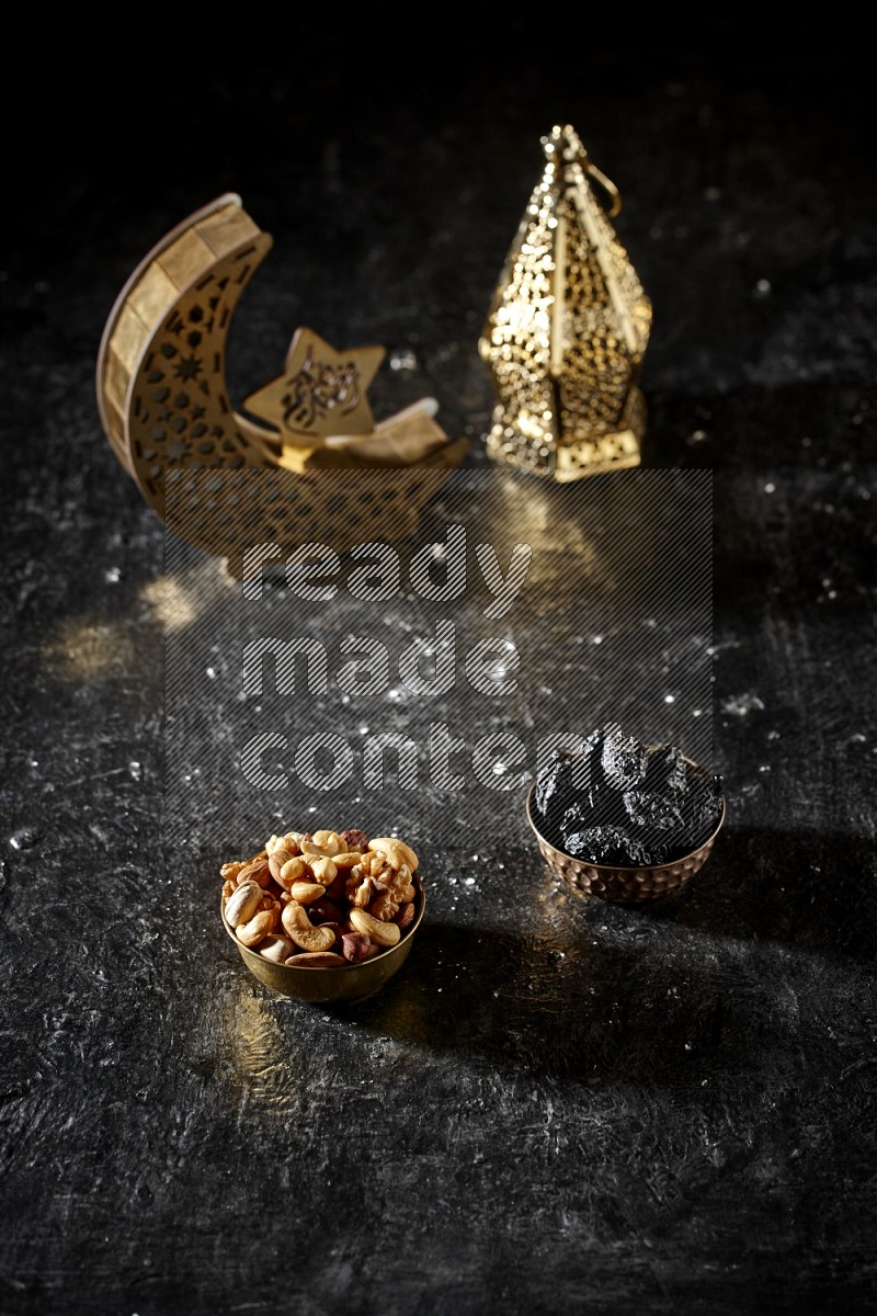 Nuts in a metal bowl with dried plums beside golden lanterns in a dark setup