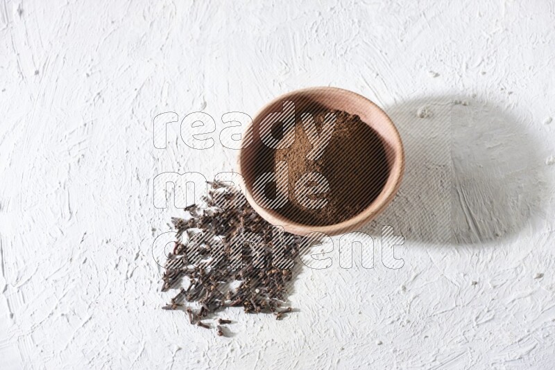A wooden bowl full of cloves powder with whole cloves beside it on a textured white flooring