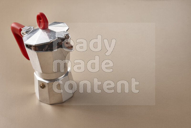 A moka pot with red handle surrounded by roasted coffee beans on beige background