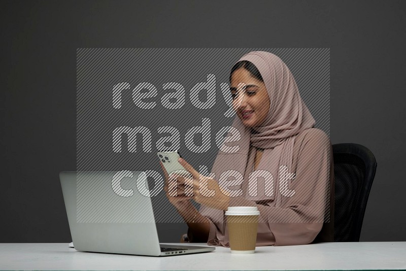 A Saudi woman Sitting on her desk Texting on a Gray Background wearing Brown Abaya with Hijab
