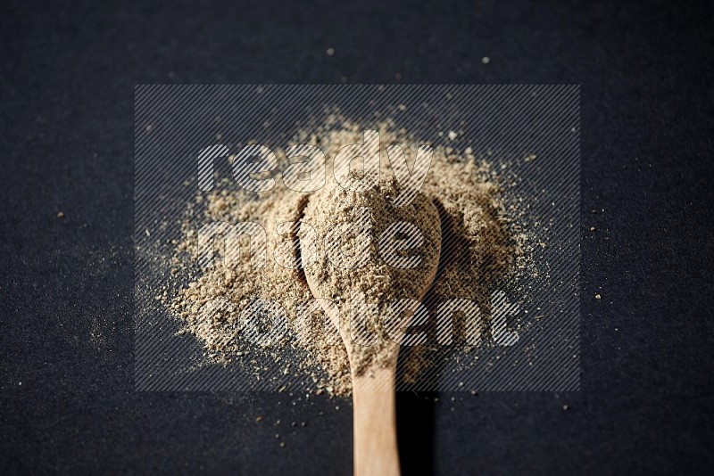A wooden spoon full of cardamom powder and powder spreaded on black flooring