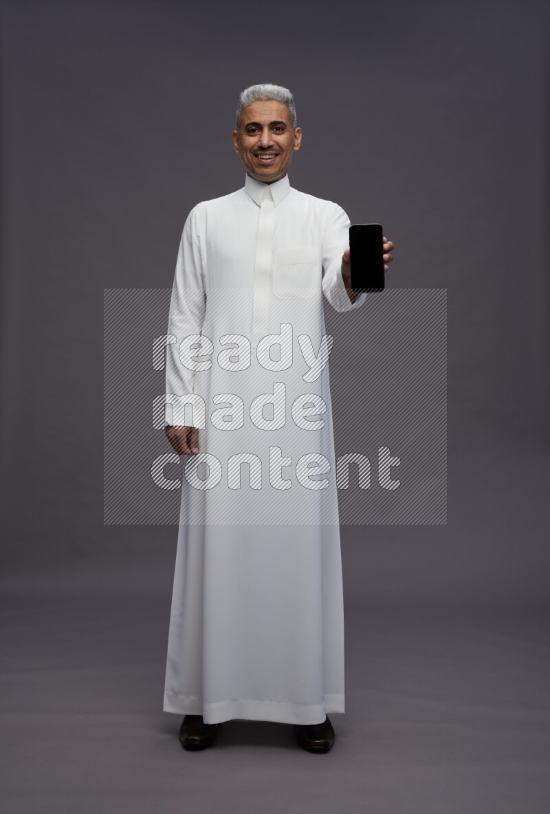 Saudi man wearing thob standing showing phone to camera on gray background