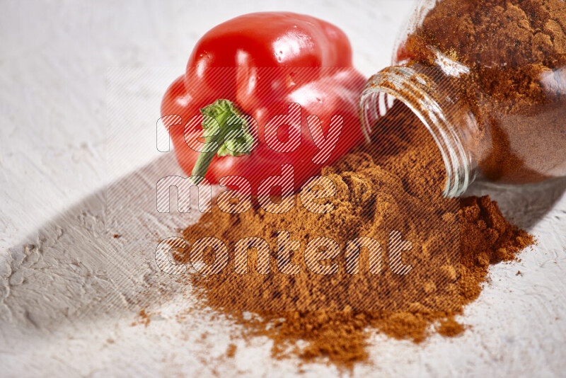 A glass jar full of ground paprika powder flipped with some spilling powder on white background