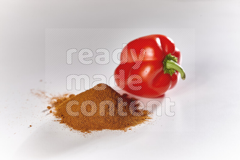 A bunch of ground paprika powder with a red bell pepper beside it on white background