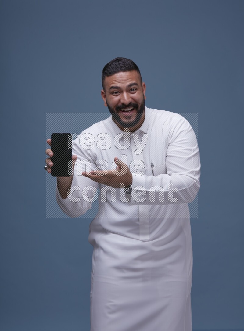 A man Showing his phone in Blue Background wearing Saudi Thob