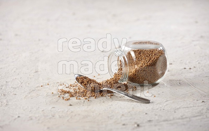 A glass spice jar and a metal spoon full of mustard seeds and jar is flipped with fallen seeds on a textured white flooring in different angles