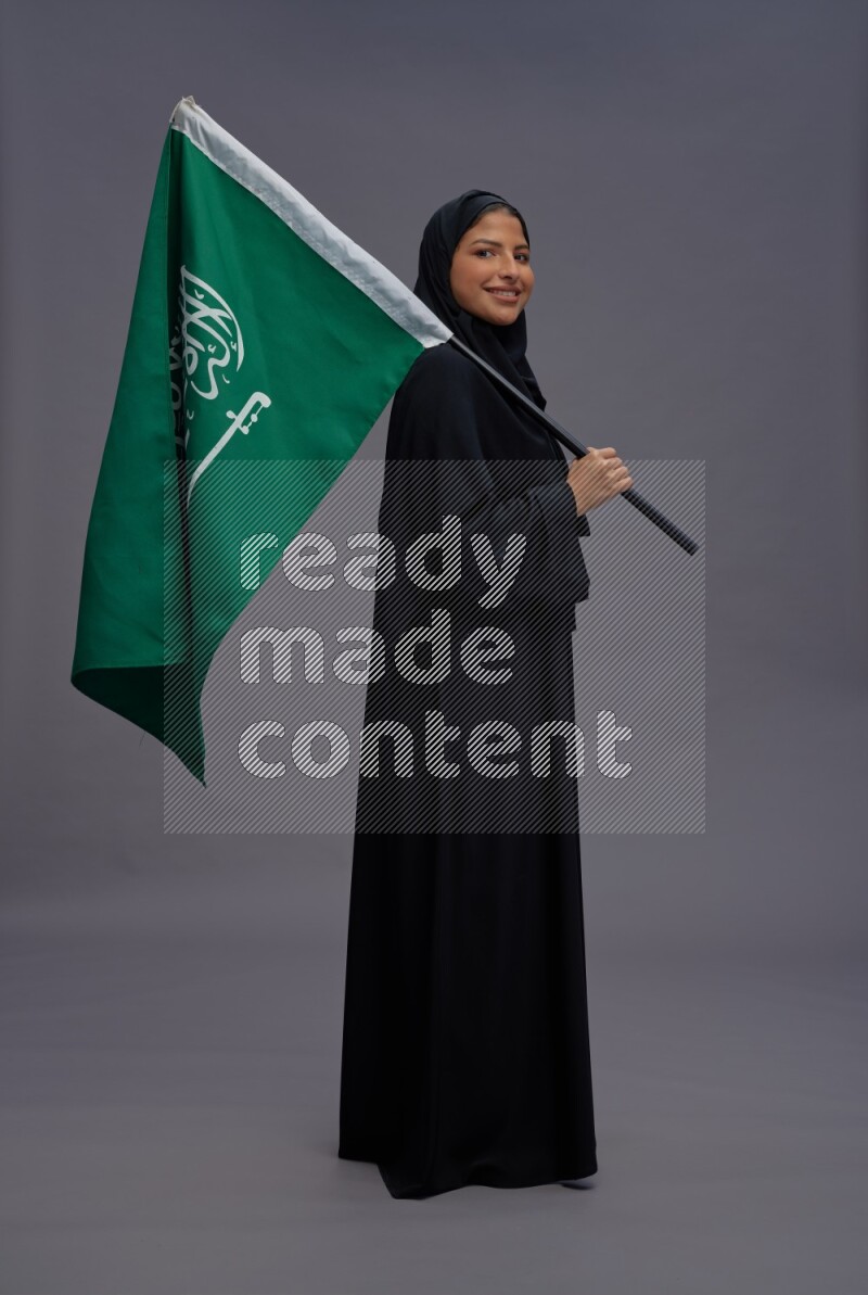 Saudi woman wearing Abaya standing holding Saudi flag on gray background