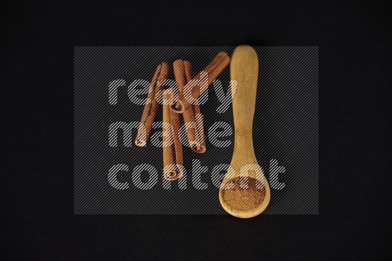 بودرة القرفة في ملعقة خشبية مع أعواد القرفة بجانبها على خلفية سوداء