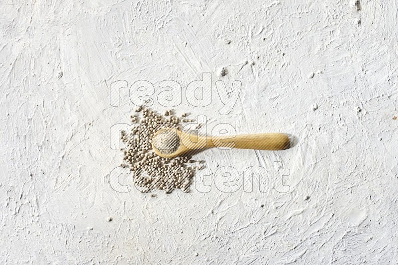 A wooden spoon full of white pepper powder and white pepper beads on textured white flooring