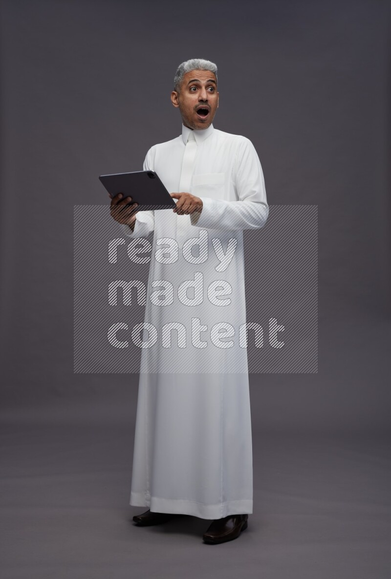 Saudi man wearing thob standing working on tablet on gray background