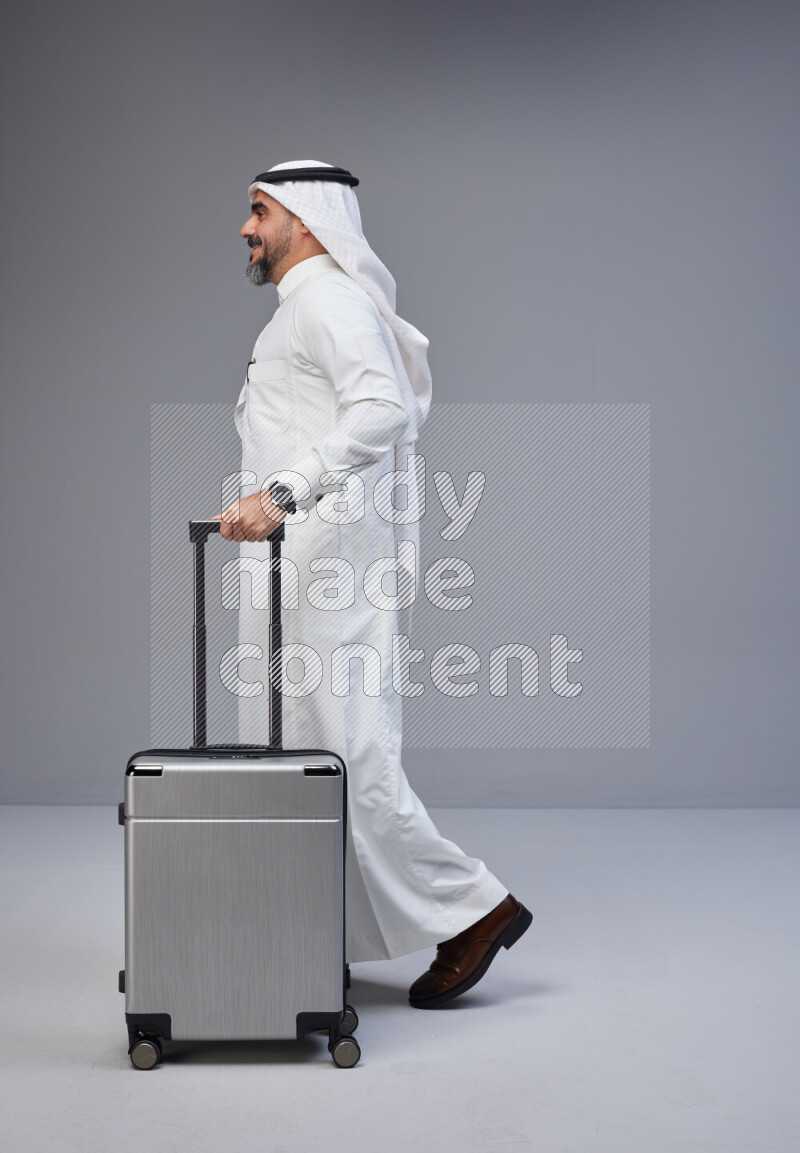 Saudi man wearing Thob and white Shomag standing holding Travel bag on Gray background