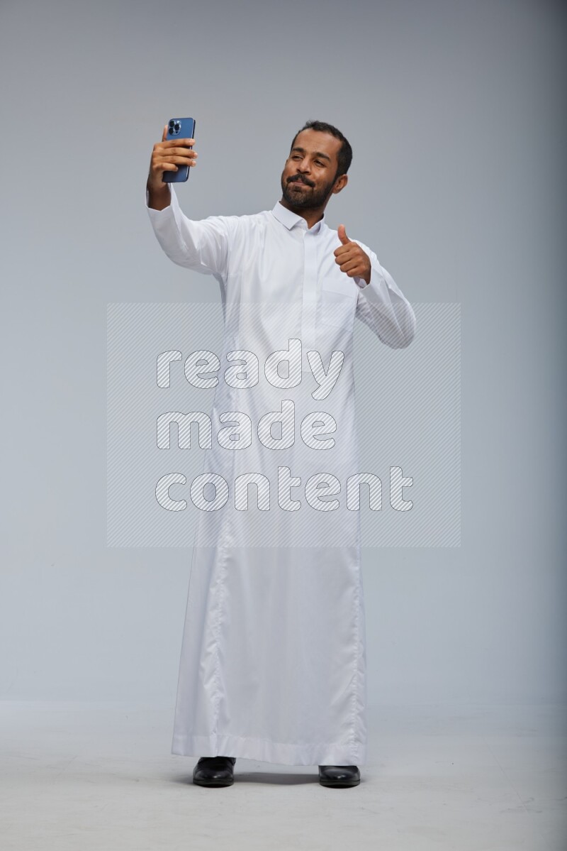 Saudi man wearing Thob standing taking selfie on Gray background