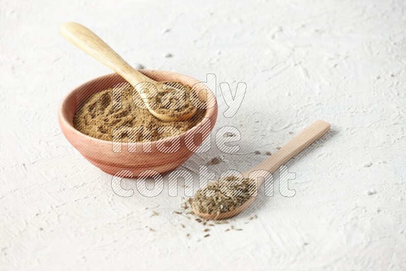 A wooden bowl and 2 wooden spoons full of cumin powder and cumin seeds on textured white flooring
