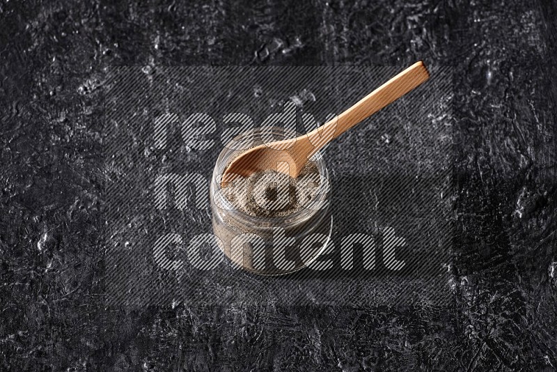 A glass jar full of black pepper powder and a wooden spoon on a textured black flooring