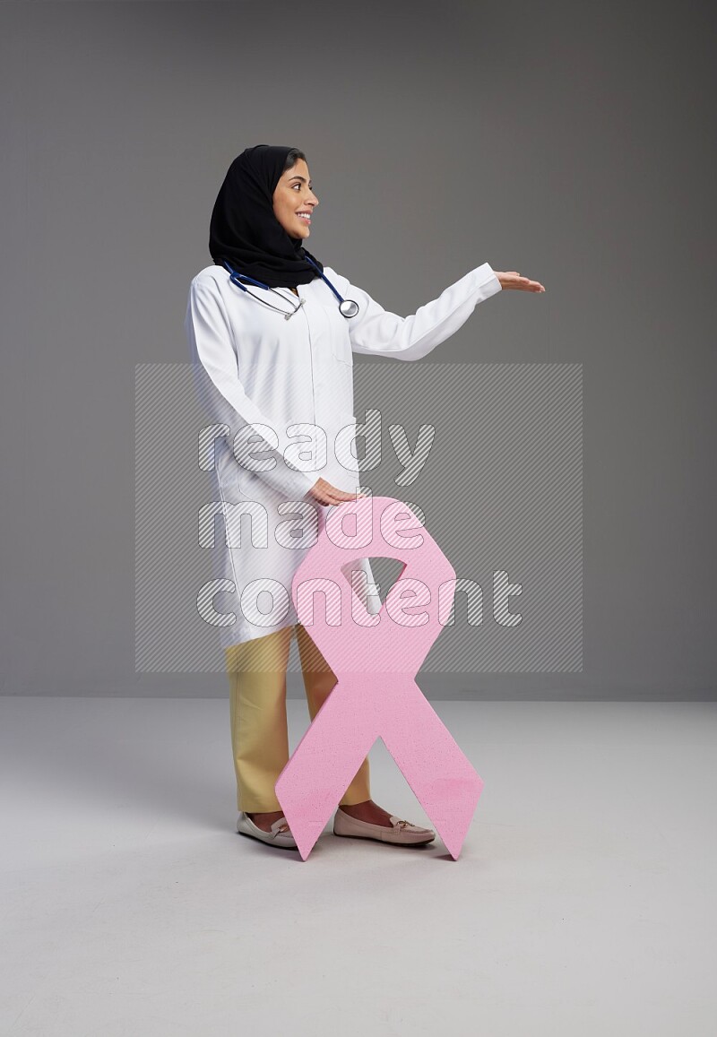 Saudi woman wearing lab coat with stethoscope standing holding awareness ribbon symbols standing on Gray background