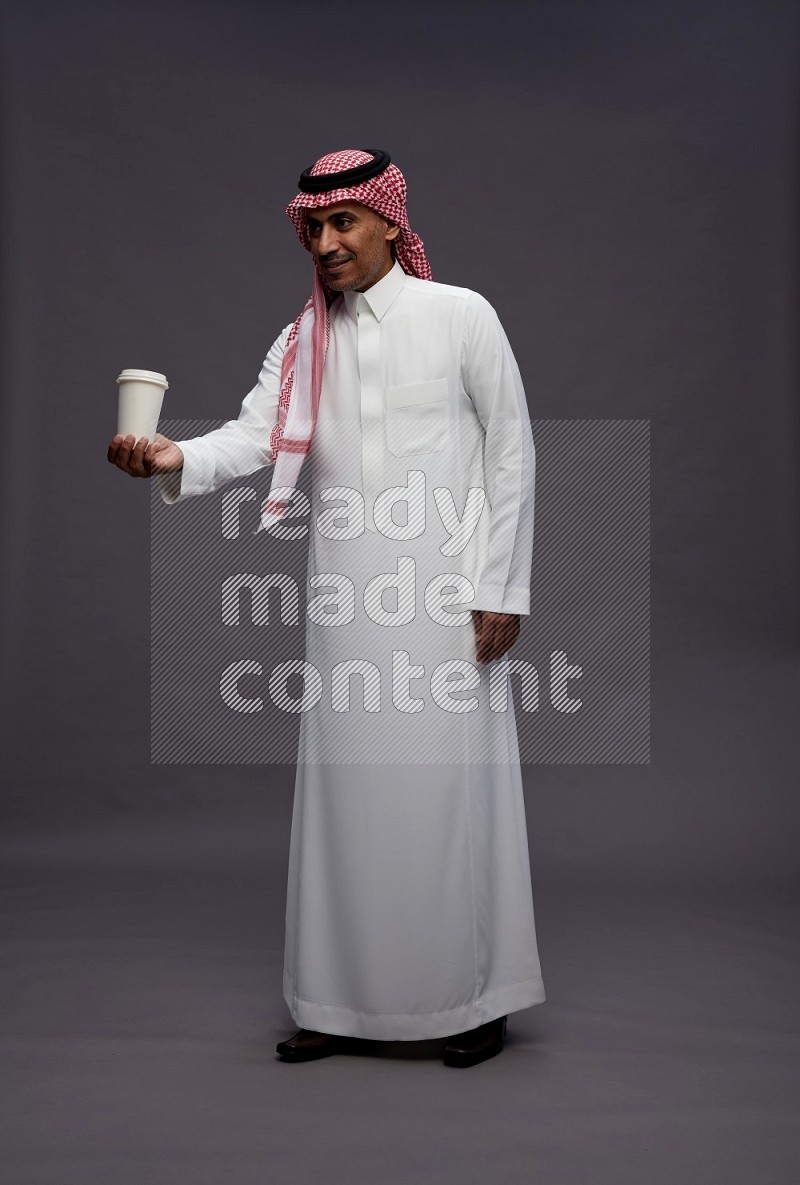 Saudi man wearing thob and shomag standing holding paper cup on gray background