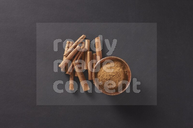 Cinnamon sticks stacked beside a wooden bowl full of cinnamon powder on black background