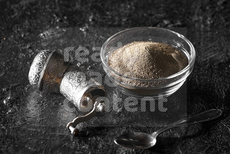 A glass bowl full of black pepper powder with a turkish metal pepper grinder and a metal spoon on textured black flooring