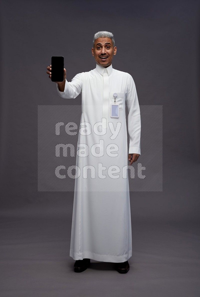 Saudi man wearing thob with pocket employee badge standing showing phone to camera on gray background