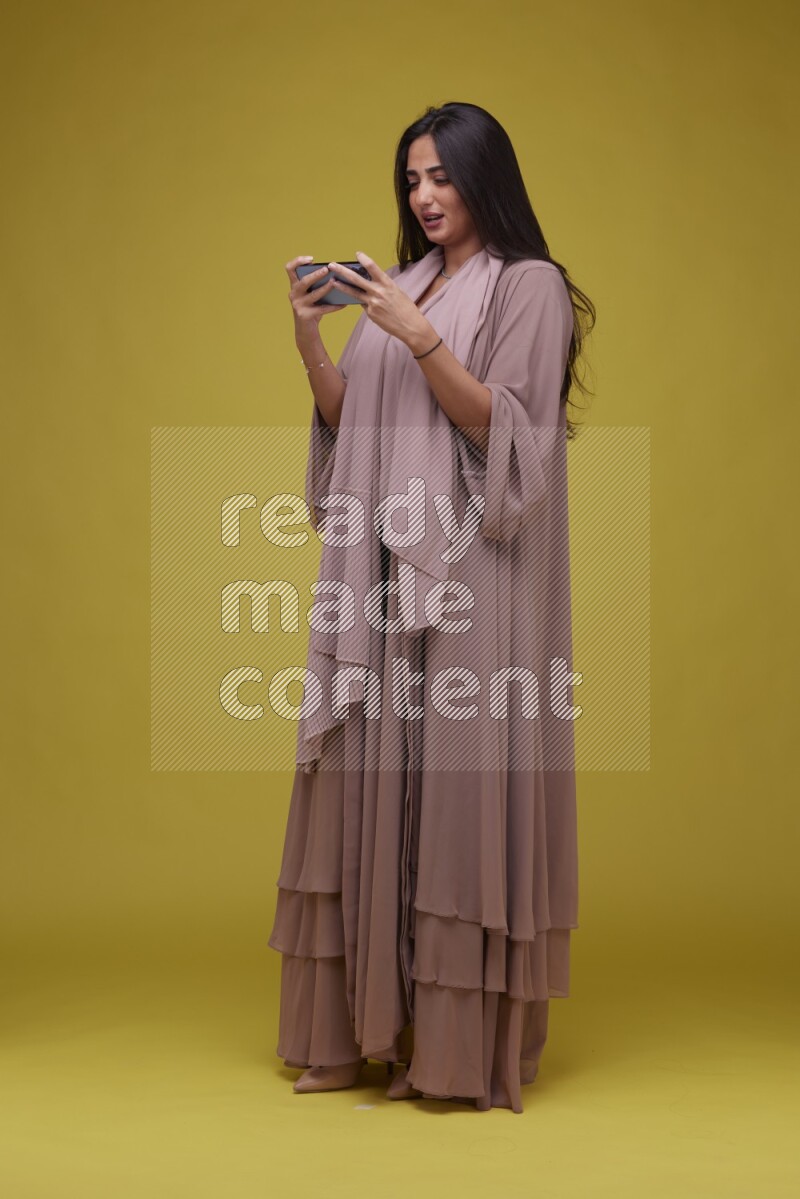A woman Playing a Game on her smartphone on a Yellow Background wearing Brown Abaya