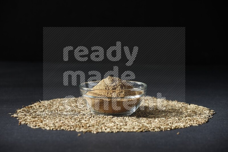 A glass bowl full of cumin powder surrounded by cumin seeds on black flooring