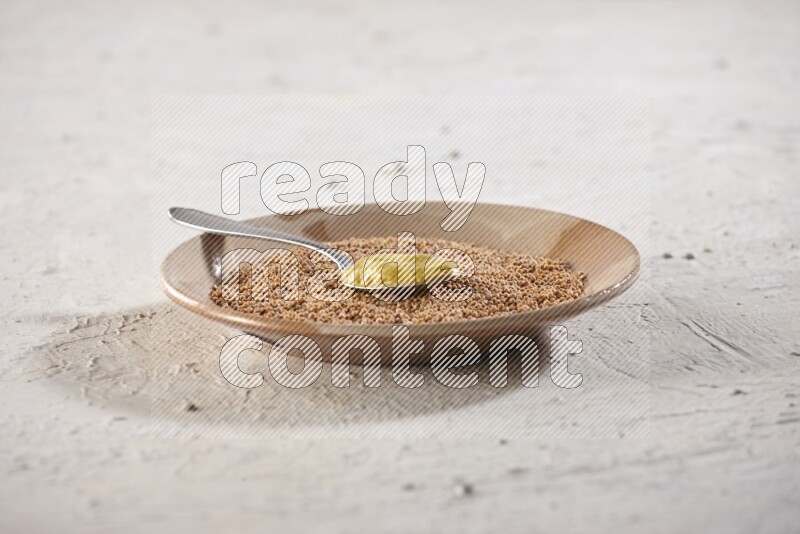 A beige pottery plate filled with mustard seeds with a metal spoon full of mustard paste in it on a textured white flooring