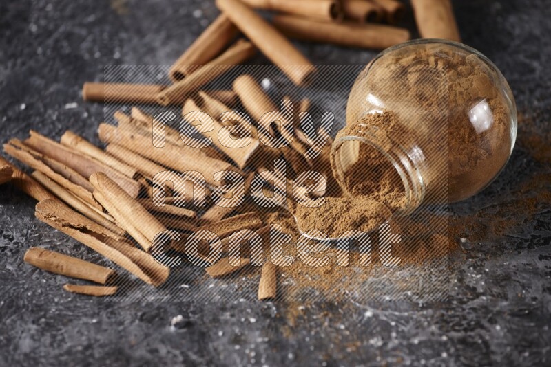 Herbal glass jar full cinnamon powder flipped and a metal spoon full of powder surrounded by cinnamon sticks on textured black background in different angles