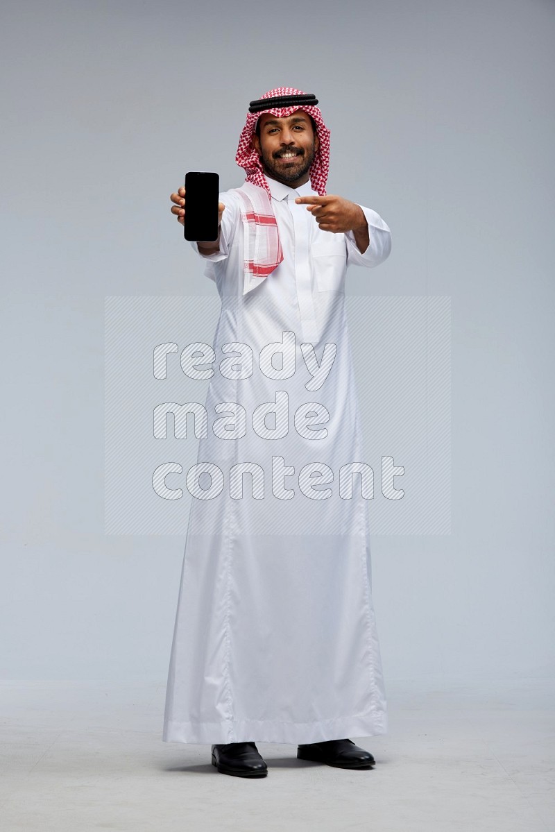 Saudi man Wearing Thob and shomag standing showing phone to camera on Gray background