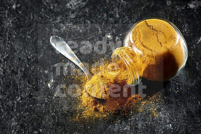 A flipped glass spice jar and a metal spoon full of turmeric powder and powder spilled out of it on textured black flooring