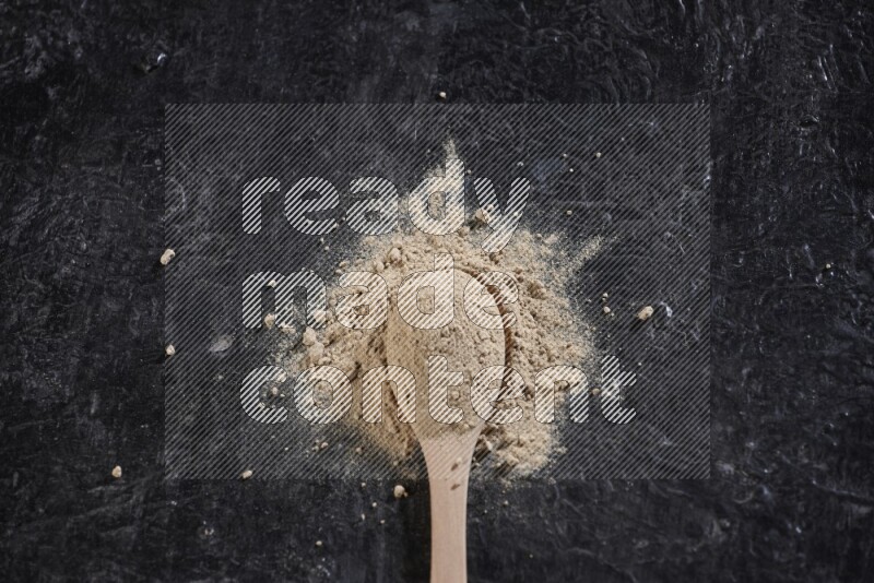 A wooden spoon full of garlic powder with scattered powder around it on a textured black flooring