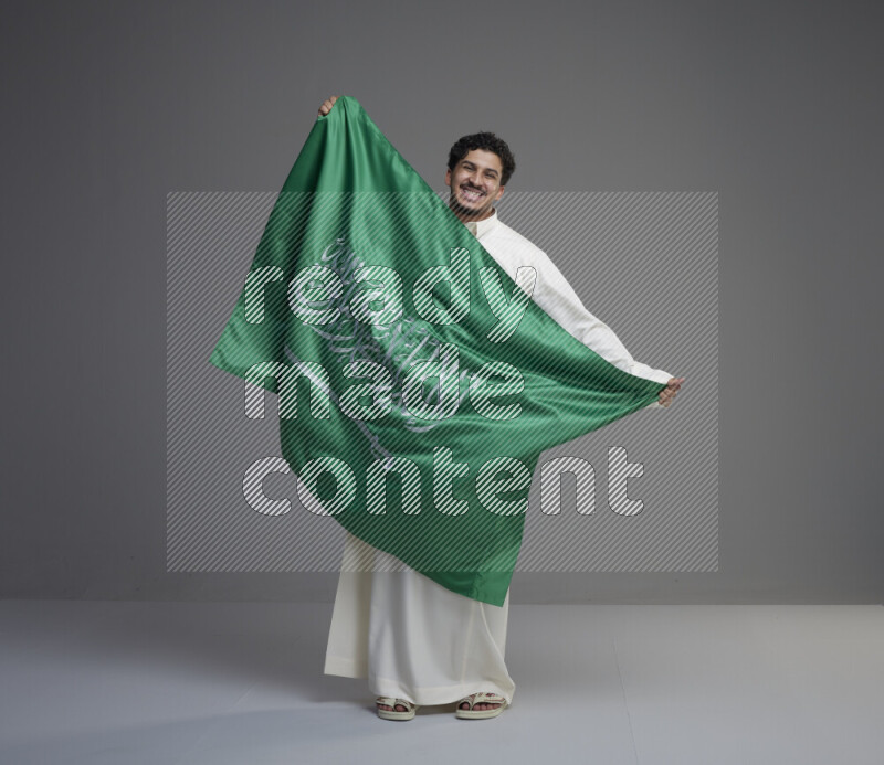 A Saudi man standing wearing thob holding big Saudi flag on gray background