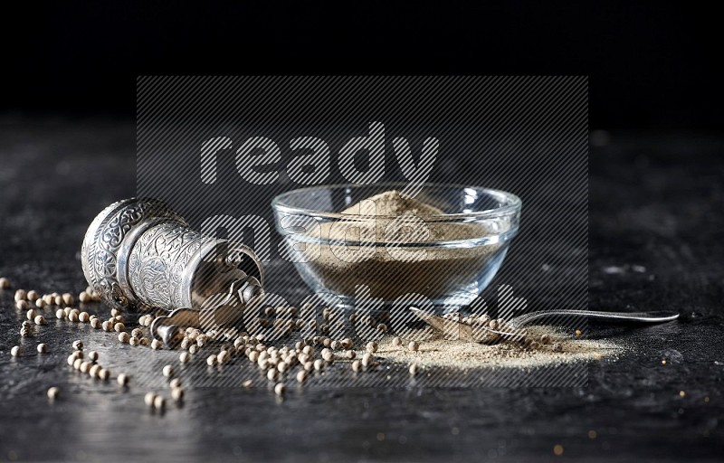 A glass bowl full of white pepper powder with pepper beads, a metal grinder and a metal spoon on textured black flooring