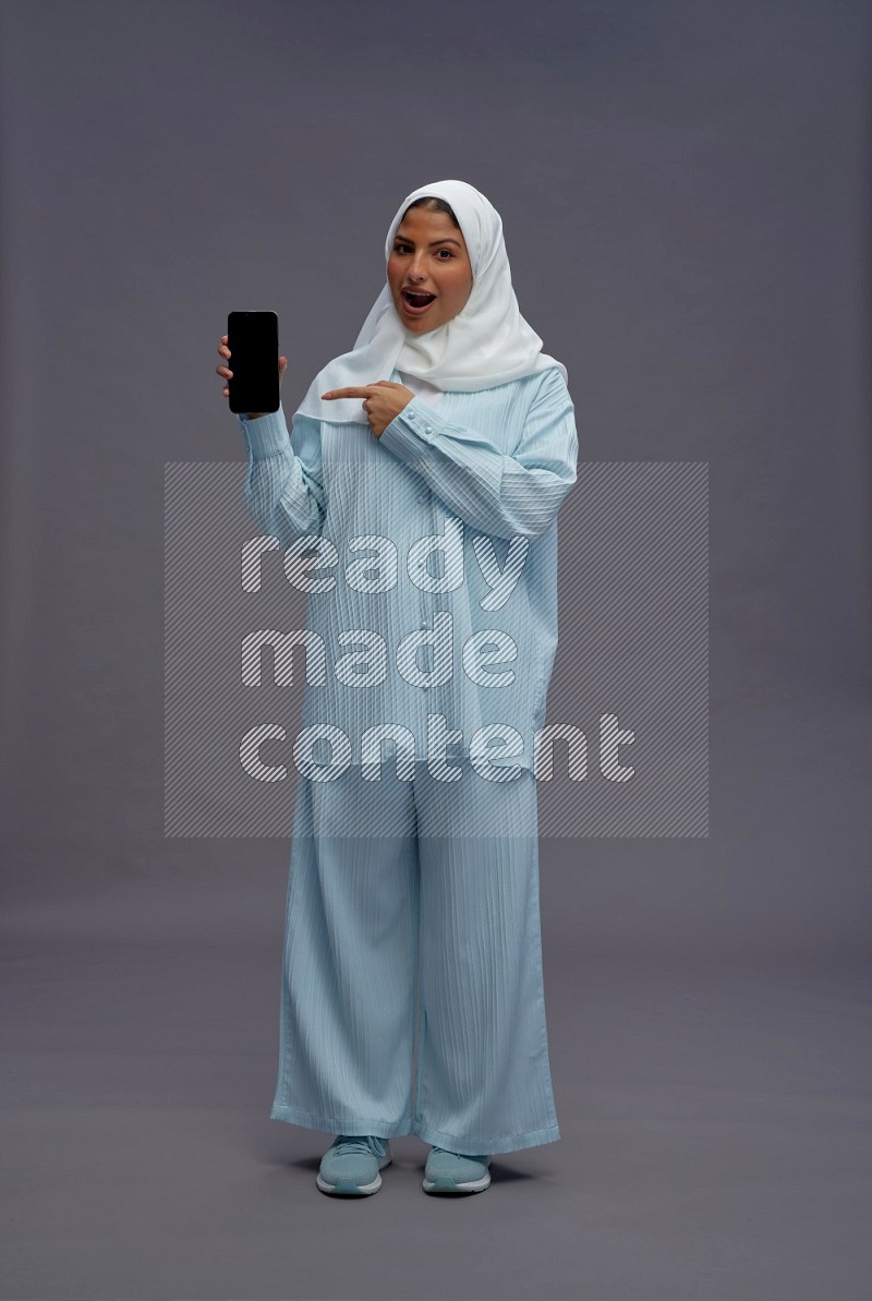 Saudi woman wearing hijab clothes standing showing phone to camera on gray background