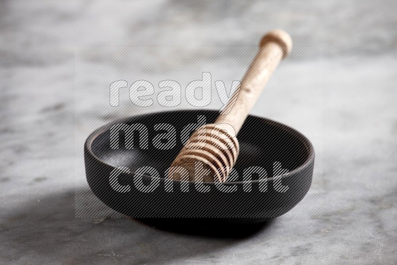 Black Pottery bowl with wooden honey handle in it, on grey marble flooring, 15 degree angle
