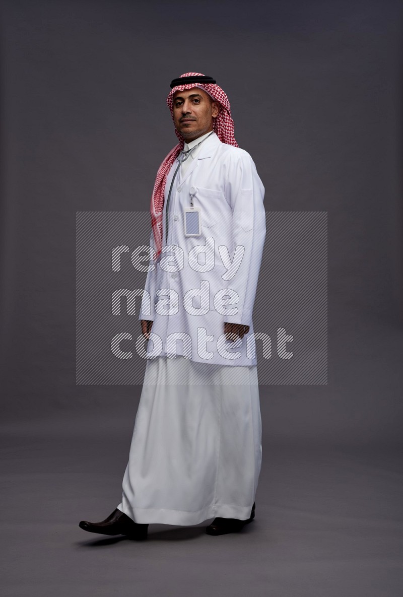 Saudi man wearing thob with lab coat and shomag with pocket employee badge with stethoscope standing interacting with the camera on gray background