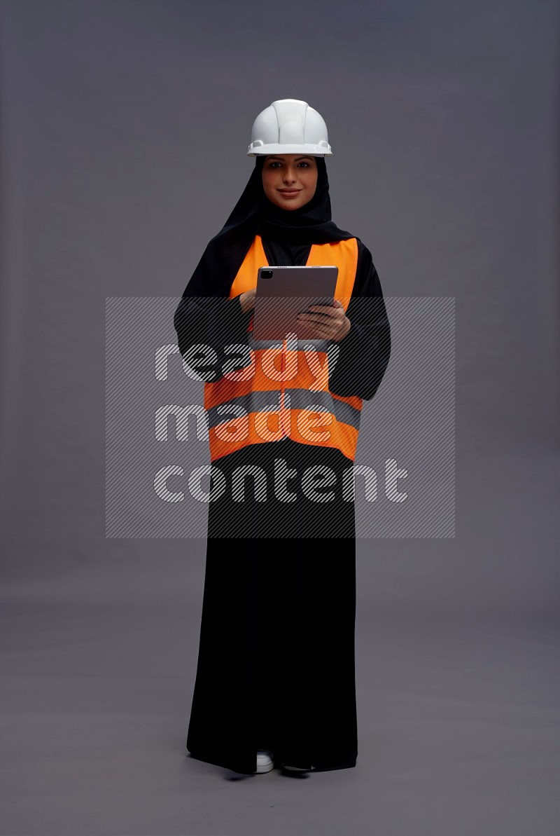 Saudi woman wearing Abaya with engineer vest standing working on tablet on gray background
