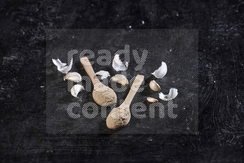 2 wooden spoons full of garlic powder with cloves and peels on a textured black flooring in different angles