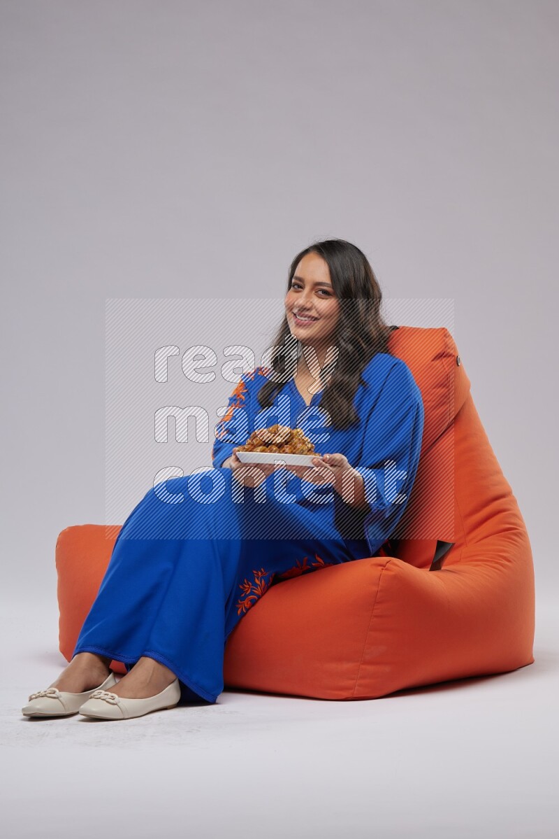 A Woman sitting on an orange beanbag wearing Jalabeya holding a plate of dates