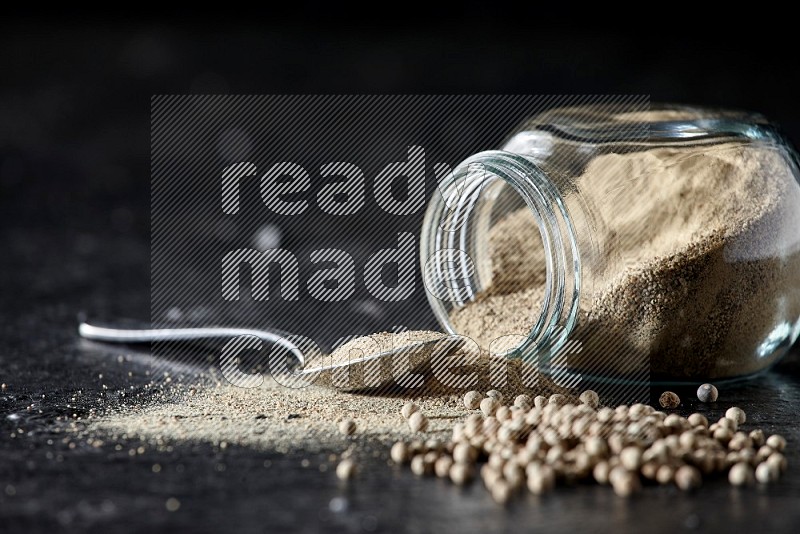 A flipped herbal glass jar and metal spoon full of white pepper powder with spilled powder and pepper beads on textured black flooring