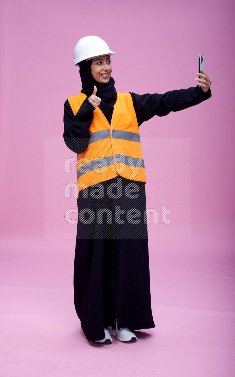 Saudi woman wearing Abaya with engineer vest and helmet standing taking selfie on pink background