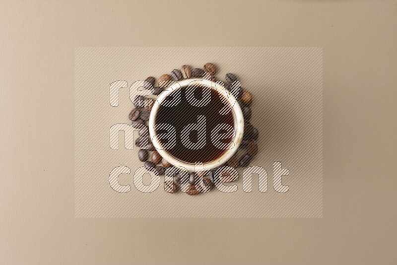 A beige pottery cup of coffee surrounded by roasted coffee beans on beige background