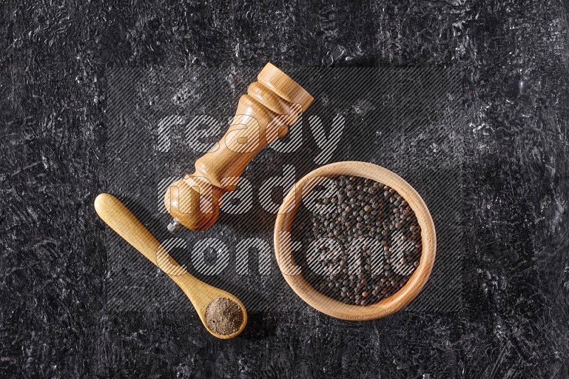 A wooden bowl full of black pepper and a wooden spoon full of black pepper powder and a wooden grinder on a textured black flooring